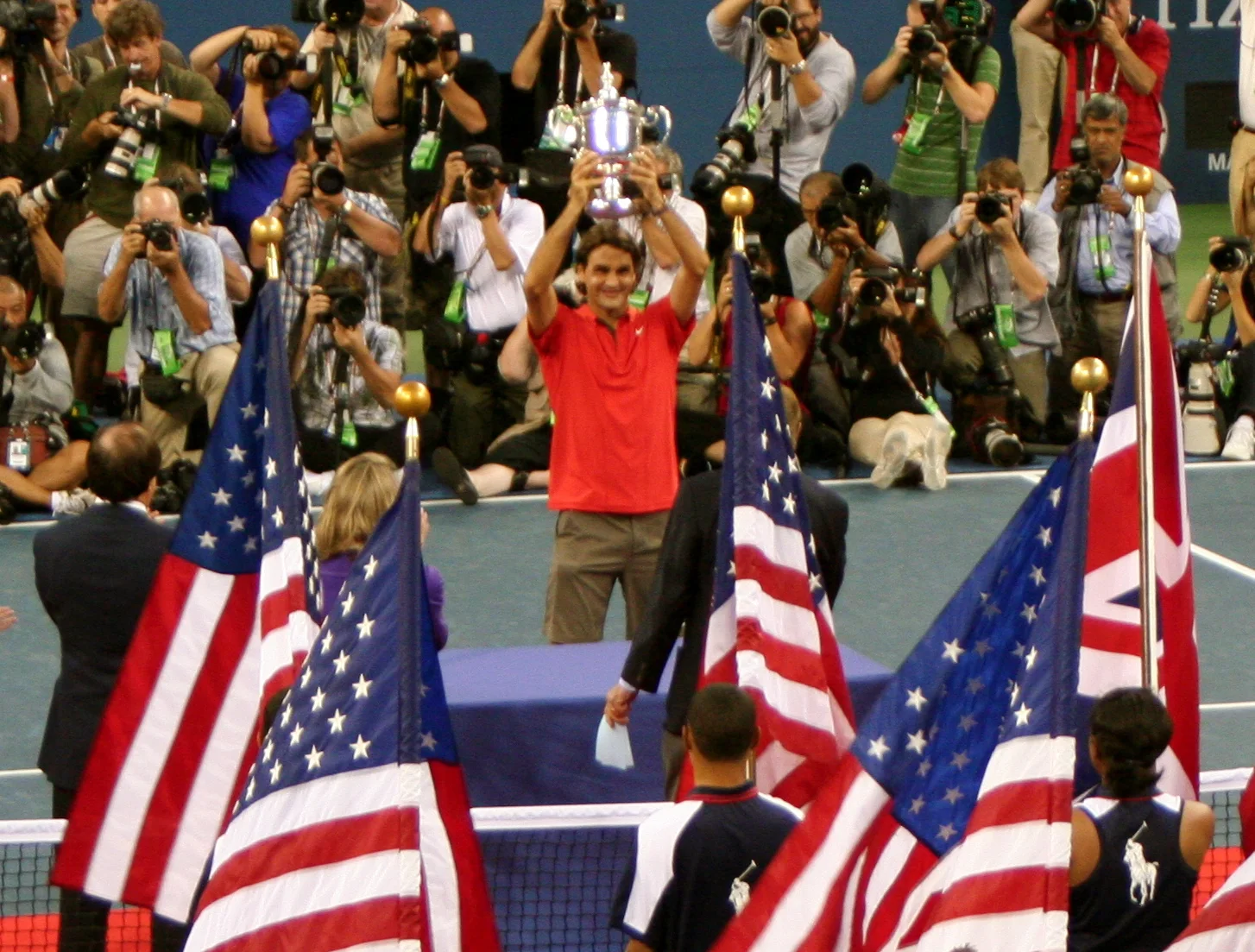 Roger Federer, de camisa vermelha, ergue o troféu do US Open de 2008 diante de bandeiras americanas e fotógrafos.